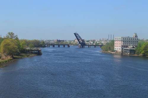 A calm river scene with a raised drawbridge, green trees, and buildings along the shore under a clear blue sky.