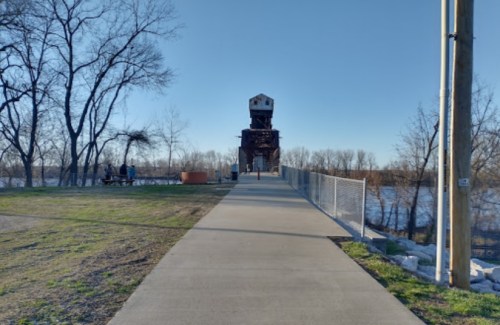 A paved path leads to a tall, old structure by a river, surrounded by trees and a fence.