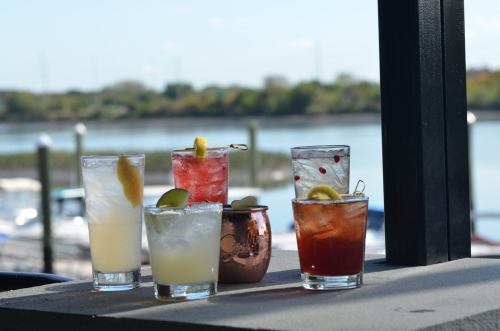 A variety of colorful cocktails on a table by the water, featuring garnishes and different glass styles.