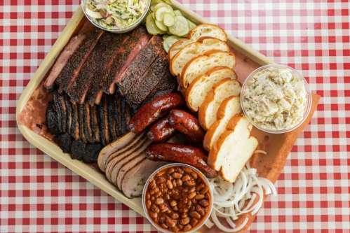A wooden tray filled with assorted barbecue meats, sliced bread, coleslaw, pickles, and baked beans on a checkered tablecloth.