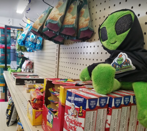 A green alien plush toy in a hoodie sits on a shelf among various snacks and treats in a store.