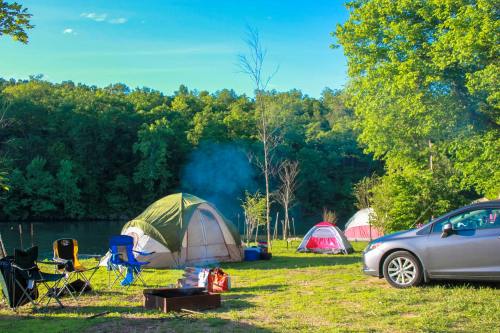 A campsite with tents, a fire pit, and a car near a river, surrounded by trees and blue skies.