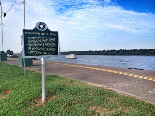 Sign about the Mississippi River Levee beside a river, with a boat in the water and green grass in the foreground.