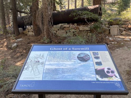 Sign titled "Ghost of a Sawmill" in a forest, with a large, rusted pipe and stone foundation in the background.