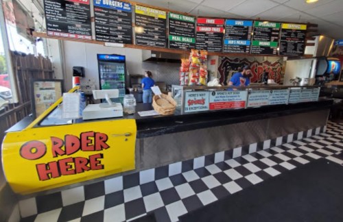 A colorful fast-food counter with a menu board, cash register, and staff preparing food in a checkered floor setting.