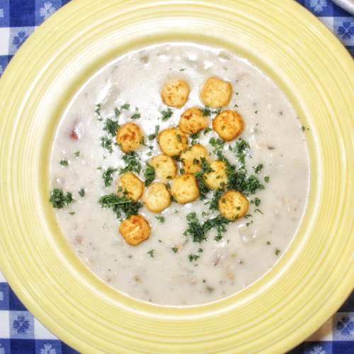 A bowl of creamy soup topped with golden croutons and sprinkled with fresh herbs, set on a blue checkered tablecloth.
