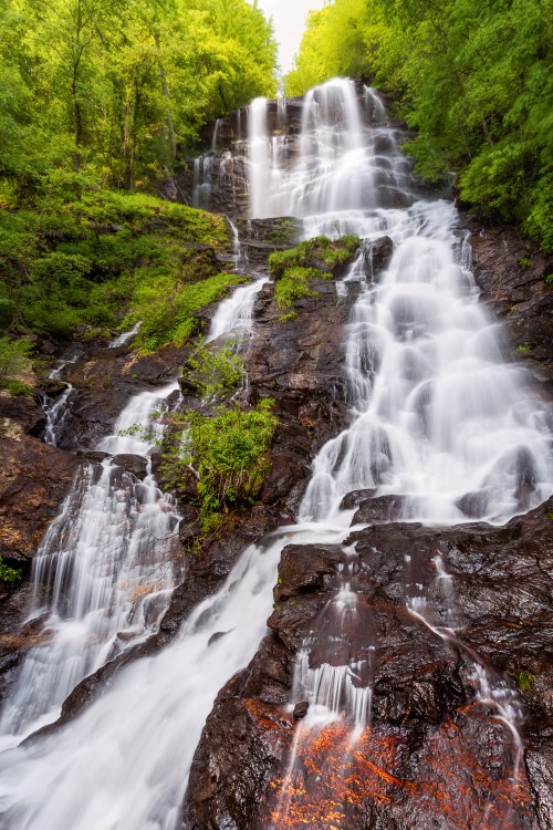 A cascading waterfall flows over rocky terrain, surrounded by lush green trees and vibrant foliage.