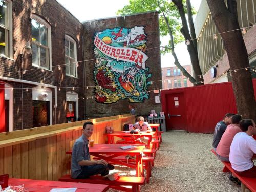 Outdoor seating area with red picnic tables, a colorful mural on the wall, and people enjoying the space.
