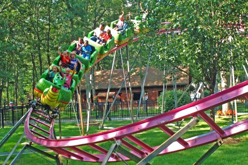 A colorful roller coaster with excited riders twists through a green park surrounded by trees and a building in the background.
