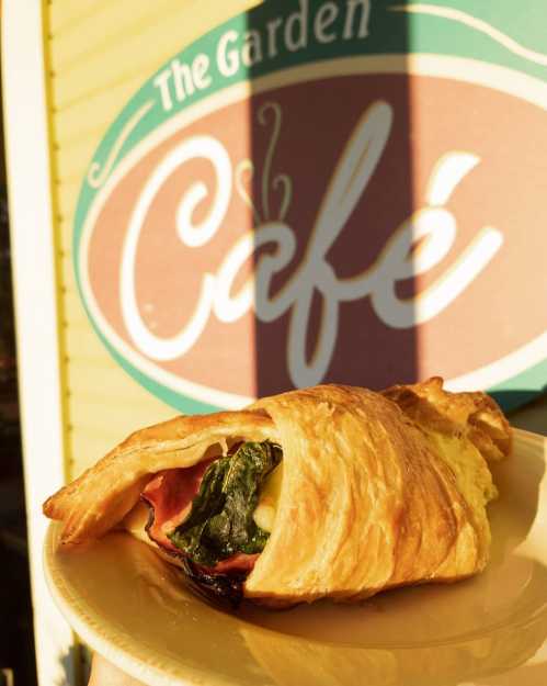 A croissant filled with spinach and ham, held in front of a café sign that reads "The Garden Café."