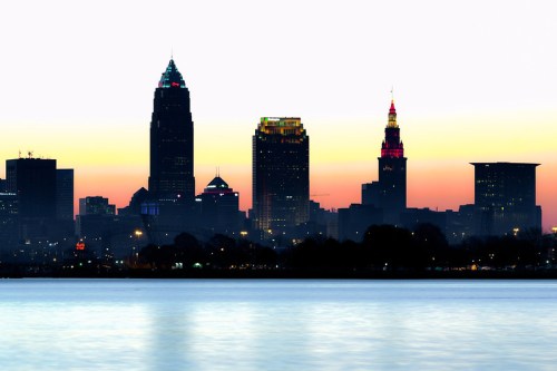 Cleveland skyline at sunset, featuring illuminated skyscrapers against a colorful sky reflected in the water.