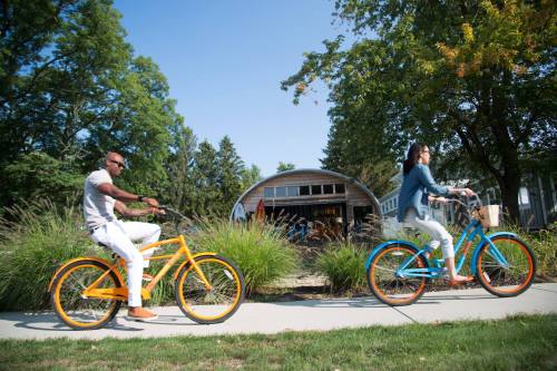 Two people ride colorful bicycles along a path in a sunny park, with greenery and a building in the background.