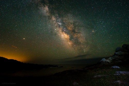 A stunning night sky filled with stars and the Milky Way, above a rocky shoreline and calm waters.
