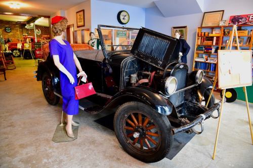 A vintage black car displayed in a museum, alongside a mannequin in a blue dress holding a red purse.