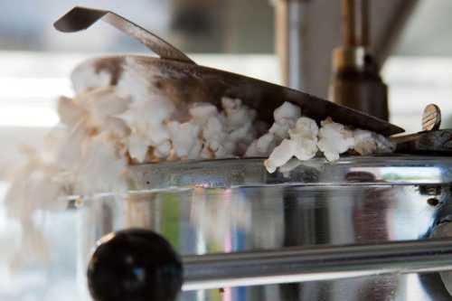 Popcorn overflowing from a metal popcorn machine, with steam rising and a scoop resting on the edge.