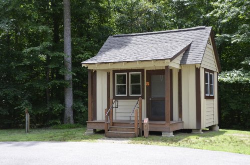 A small, charming cabin with a sloped roof, surrounded by trees and grass, featuring a porch and steps.