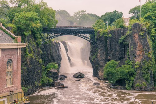 A scenic view of a waterfall cascading under a bridge, surrounded by lush greenery and rocky cliffs.