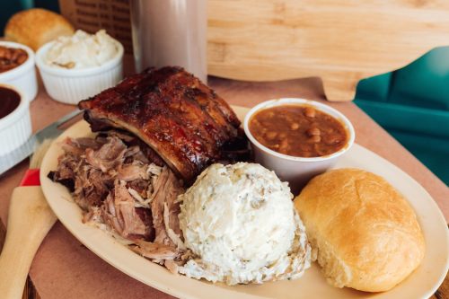 A plate of barbecue featuring pulled pork, ribs, mashed potatoes, baked beans, and a roll.