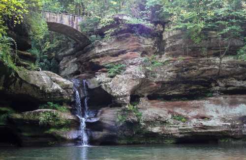 A serene waterfall cascades over rocky cliffs, surrounded by lush greenery and a stone bridge above.