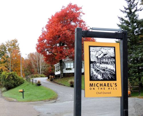 Sign for "Michael's on the Hill" with a scenic autumn backdrop of colorful trees and a winding road.