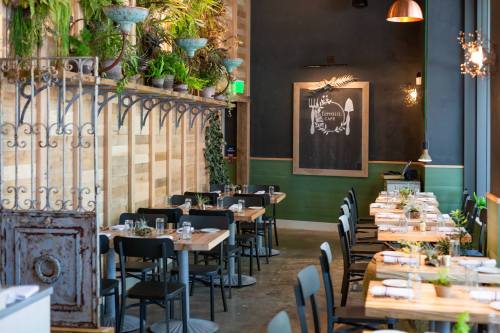 A modern restaurant interior with wooden tables, black chairs, and greenery on the walls, featuring a chalkboard menu.