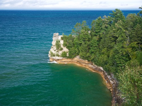 A scenic view of a rocky shoreline with lush green trees and calm blue waters under a cloudy sky.