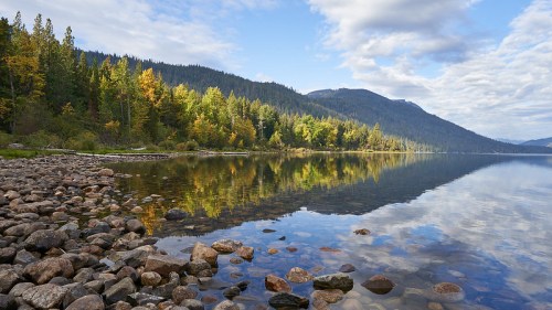 A serene lake surrounded by trees, with rocky shores and a reflection of clouds and mountains in the water.