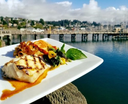 A plate of grilled fish with vegetables, set against a scenic waterfront view with boats and clouds.