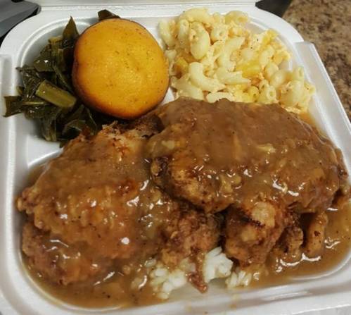 A takeout container with fried chicken smothered in gravy, rice, macaroni and cheese, collard greens, and cornbread.