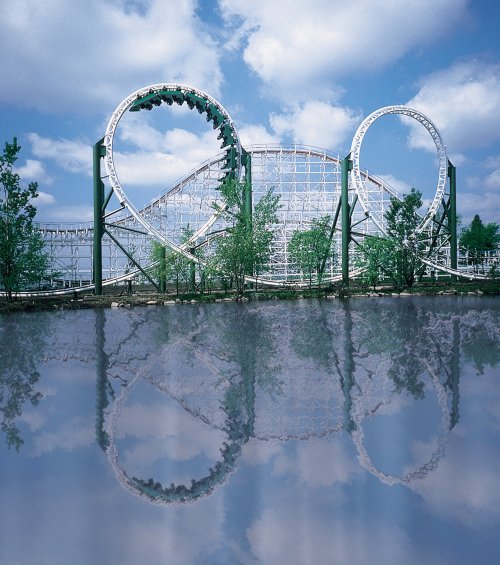 A roller coaster with two loops reflected in calm water, surrounded by trees and under a blue sky with clouds.