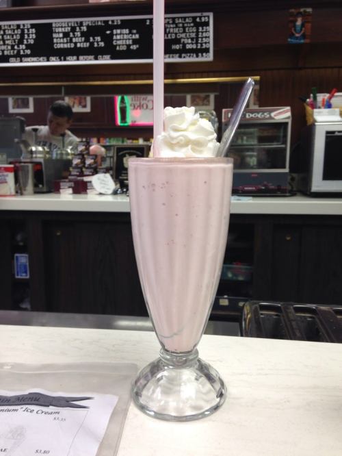 A tall glass of pink milkshake topped with whipped cream and a straw, sitting on a counter with a menu in the foreground.