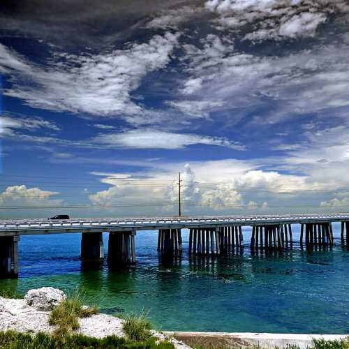 A bridge spans over calm water, with lush greenery and a cloudy blue sky in the background.