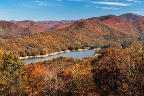 Scenic view of a lake surrounded by colorful autumn foliage and rolling mountains under a clear blue sky.