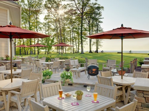 Outdoor patio with wooden tables, red umbrellas, and a fire pit, surrounded by trees and a view of the lake.