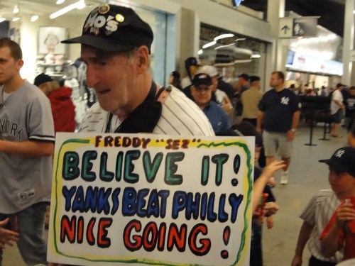 An older man in a Yankees cap holds a colorful sign celebrating the Yankees' victory over the Phillies.