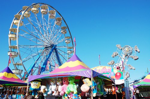 A vibrant carnival scene featuring a Ferris wheel, colorful tents, and plush toys on display under a clear blue sky.