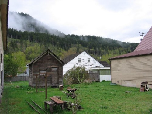 A grassy yard with wooden structures, surrounded by trees and misty mountains in the background.