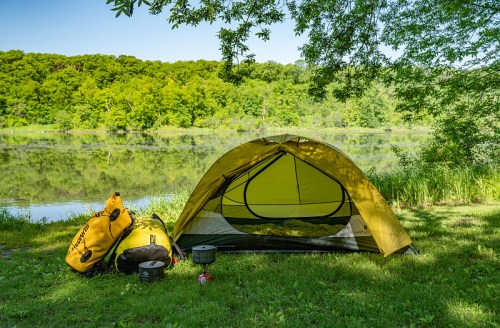 A yellow tent set up by a calm lake, surrounded by lush greenery and camping gear nearby.