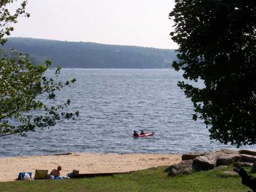 A serene lake scene with a sandy beach, a kayak on the water, and trees framing the view.