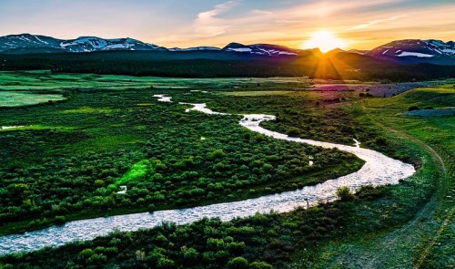 A winding river flows through a lush green landscape, with mountains and a vibrant sunset in the background.