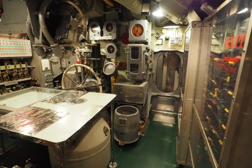 Interior of a submarine control room, featuring dials, gauges, and a large steering wheel.