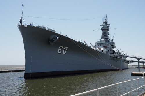 A large battleship docked at a pier, with the number 60 displayed on its bow against a clear blue sky.