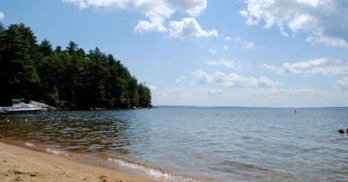 A serene beach scene with sandy shore, calm water, and trees under a partly cloudy sky.