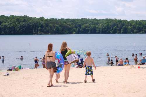 A group of people walks along the beach towards a lake, with others swimming and relaxing in the background.