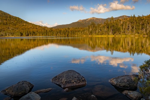 A serene lake reflects mountains and trees under a blue sky with fluffy clouds, framed by rocks in the foreground.