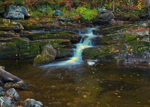 A serene waterfall cascades over moss-covered rocks into a calm pool, surrounded by autumn foliage.