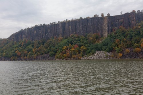 A rocky cliff with colorful autumn foliage rises above a calm river under a cloudy sky.