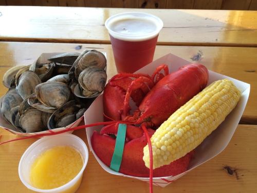 A seafood platter featuring lobster, clams, corn on the cob, and a cup of beer on a wooden table.