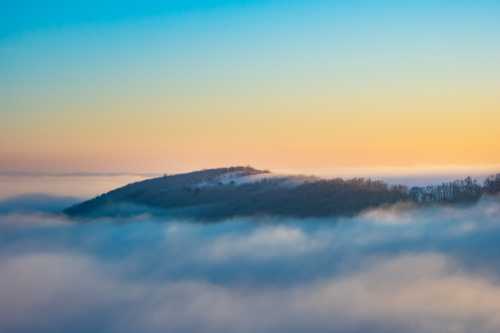 A serene landscape of a hill emerging from a sea of fog at sunrise, with a gradient sky transitioning from blue to orange.