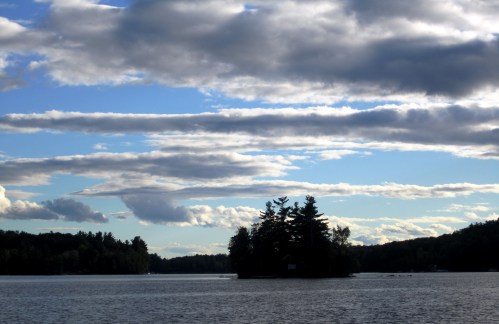 A serene lake scene with an island and trees under a sky filled with fluffy clouds.